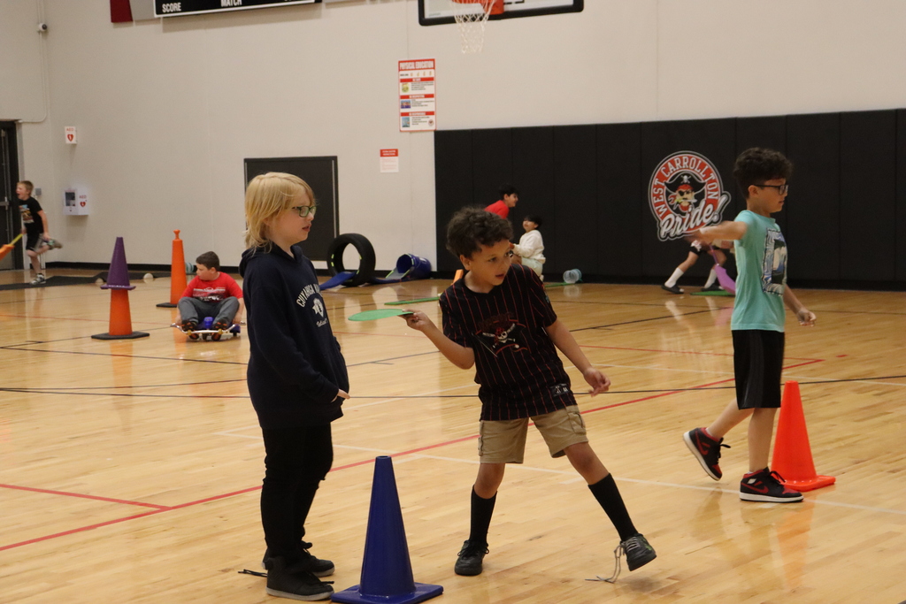 A basketball court with a boy running, another boy and a girl standing near cones, and a wall with a sign.