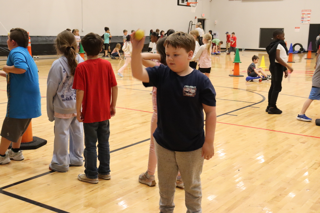 Kids inside a gym, a boy holds a ball, while others watch, with cones and a basketball hoop in the background.