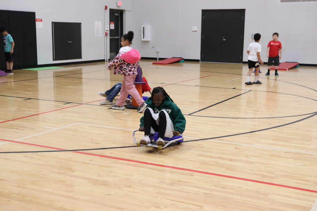Children play on a gym floor. One girl uses a skateboard while another child holds a ball.