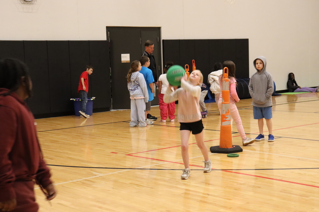 A group of children playing ball in an indoor court. A girl throws the ball in the air.