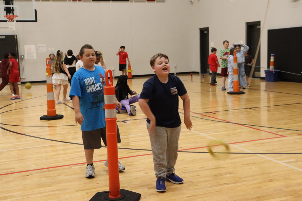 Two boys play with a ball in an indoor court. One throws it; the other catches. Orange cones and other kids are in the background.
