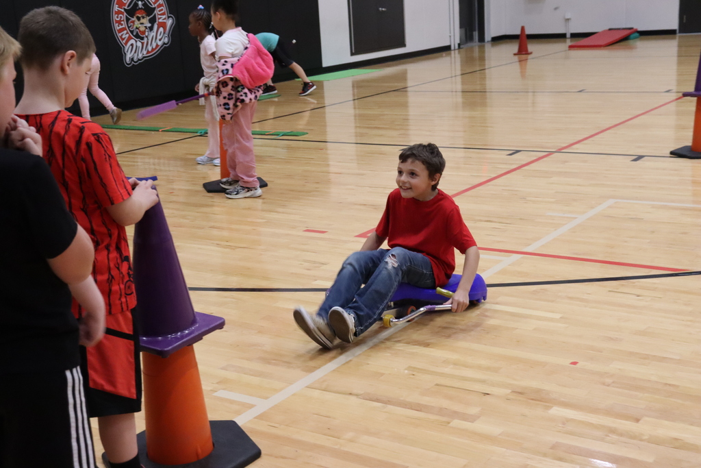 A boy rides a wheeled sled on a wooden gym floor, with traffic cones and onlookers.