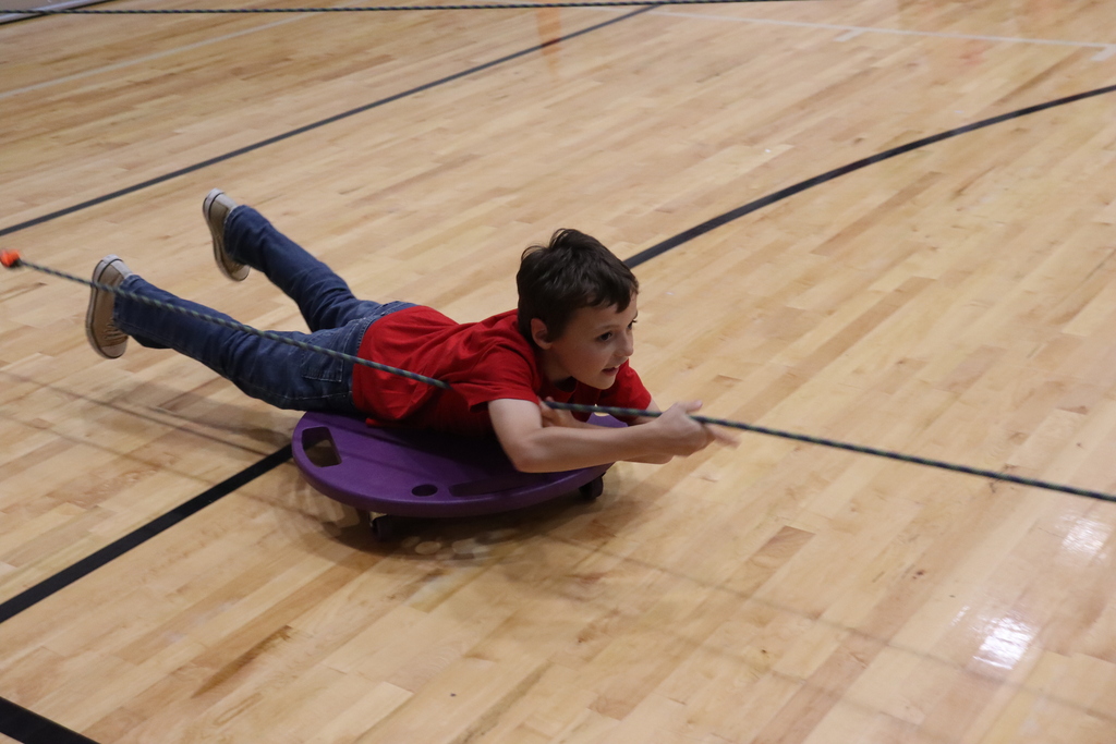 Child lying on purple sled holding black rope while lying on gym floor with black markings.