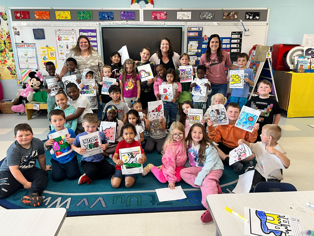 Group of children in a classroom, seated and standing, holding up drawings. Behind them are walls with various posters and displays.