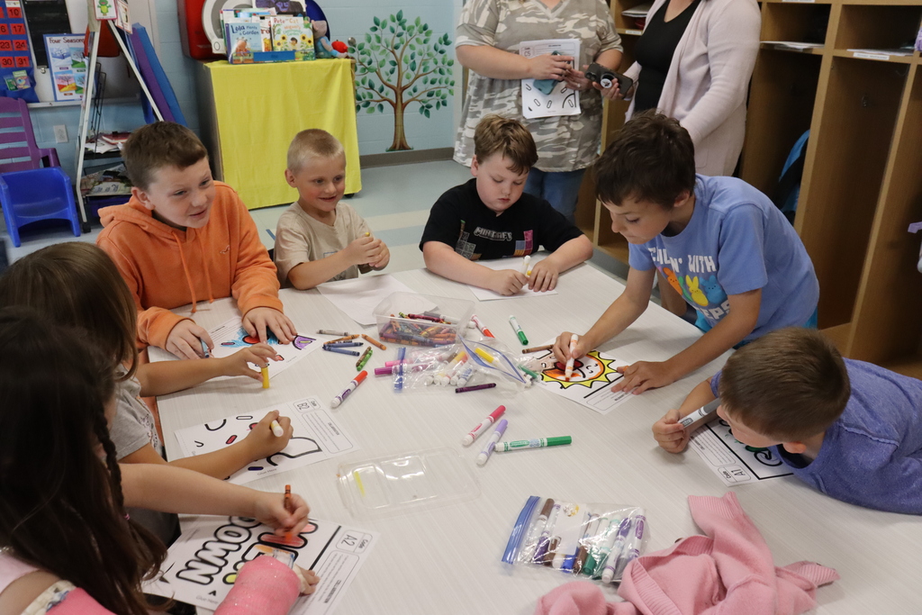 Seven children are seated around a table with art supplies. Two adults stand behind them.