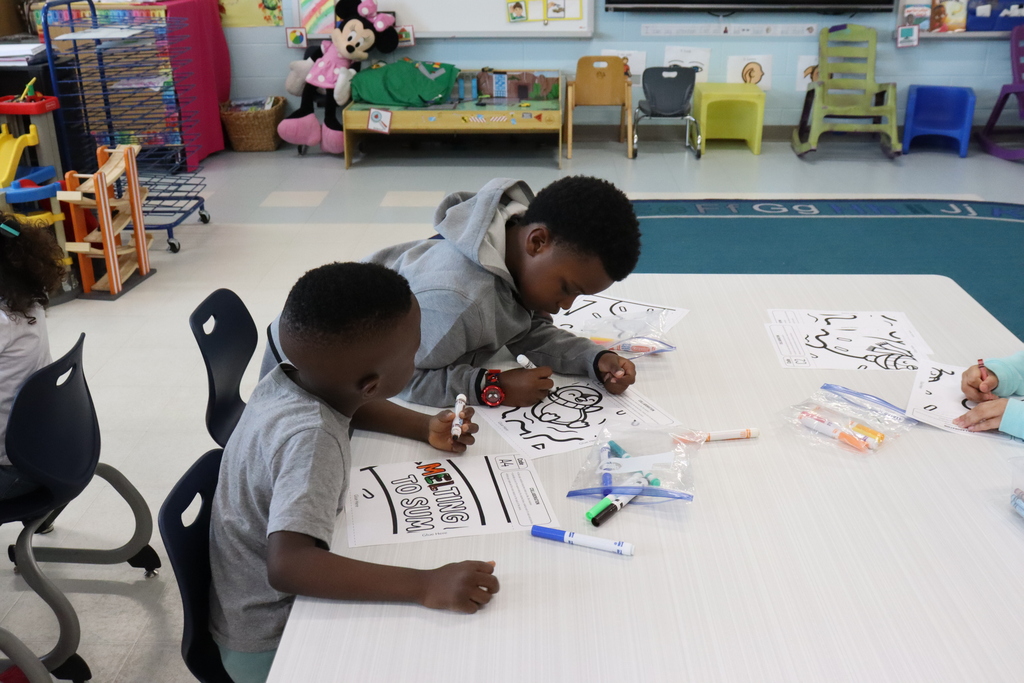Two children sit at a table, engaged in coloring and drawing activities. Various markers and papers are spread out on the table.
