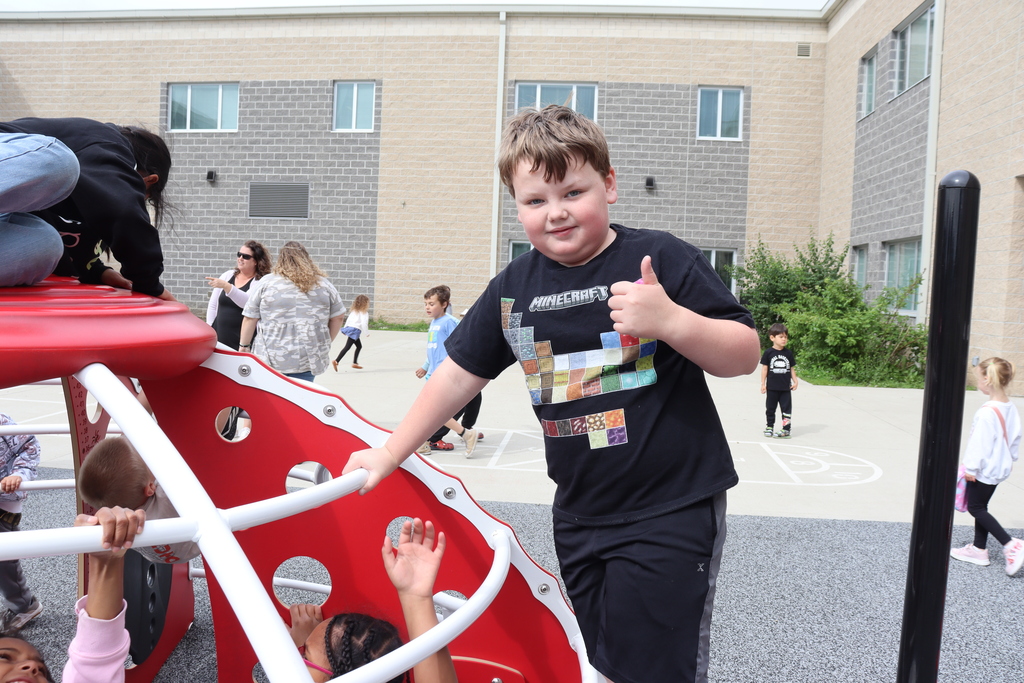 Children play on playground equipment. One boy smiles and gives a thumbs-up. Behind him is a building.