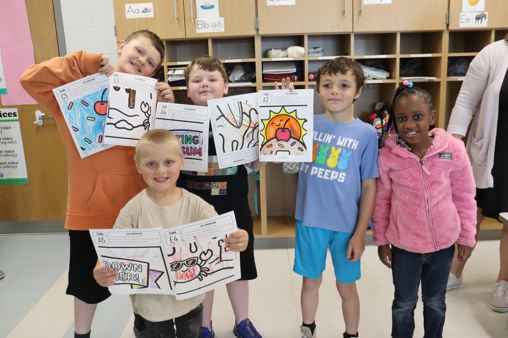 Five children, all smiling, stand in a classroom. They hold up pieces of paper with drawings. Behind them are shelves with various items.