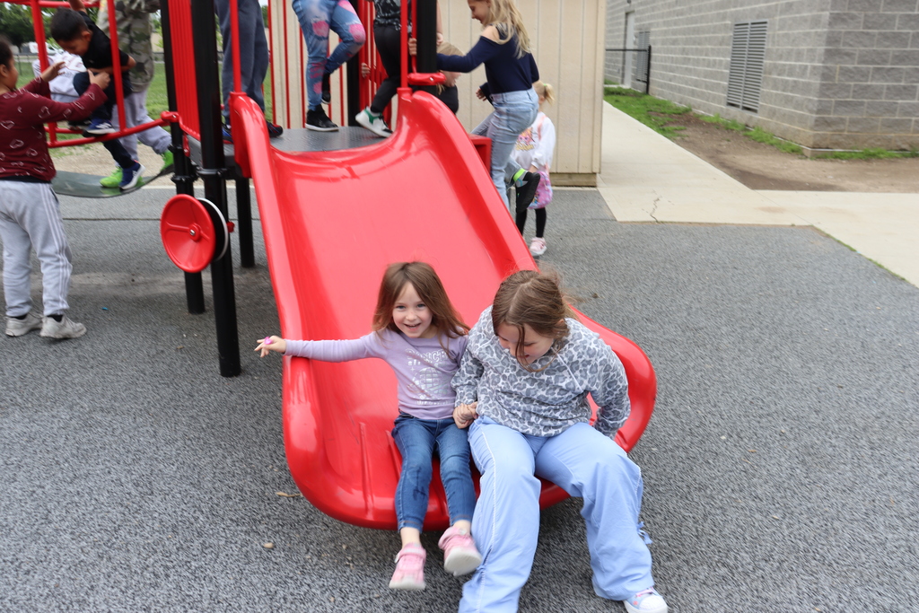Young girls slide down a red slide while a girl waits to go next. Other children play on equipment.