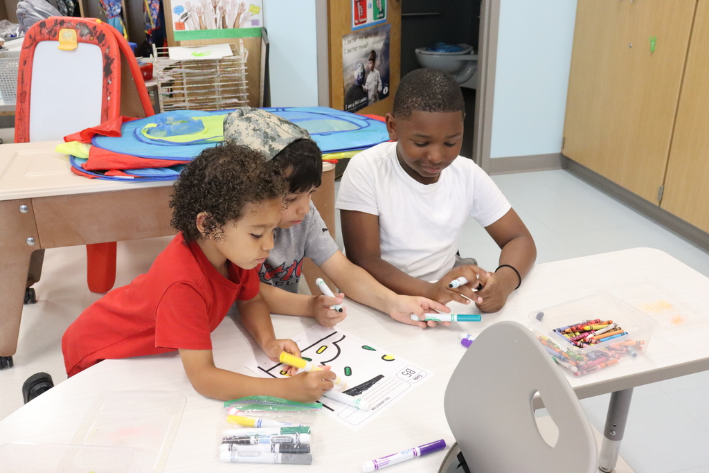 Three children seated around a table are engaged in an activity using markers and papers.