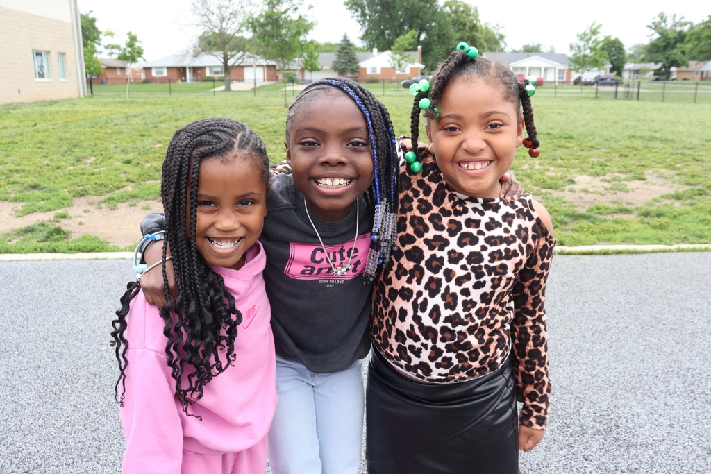 Three young girls stand together on a playground, smiling. Behind them, a grassy field stretches to a fence.