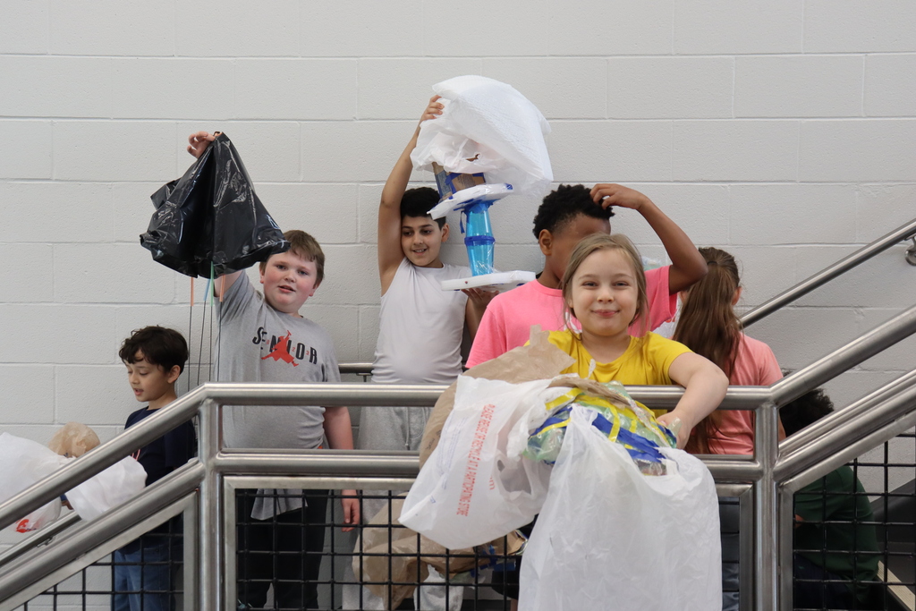 Kids holding bags of collected items on stairs, some holding up their finds. Background is a white wall.