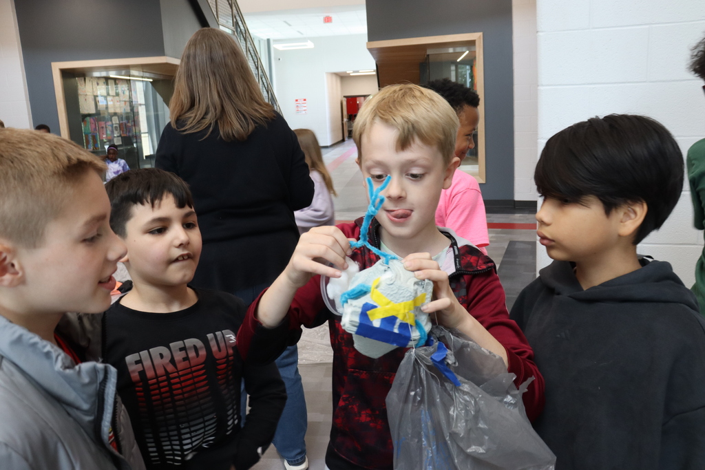 Four children look at a plastic bag held by a boy. Behind them, a woman walks. A staircase is visible on the left.