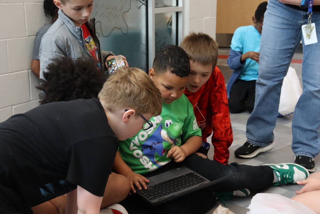 A group of children sit on the floor using a laptop, some wearing glasses and sneakers. An adult stands nearby.