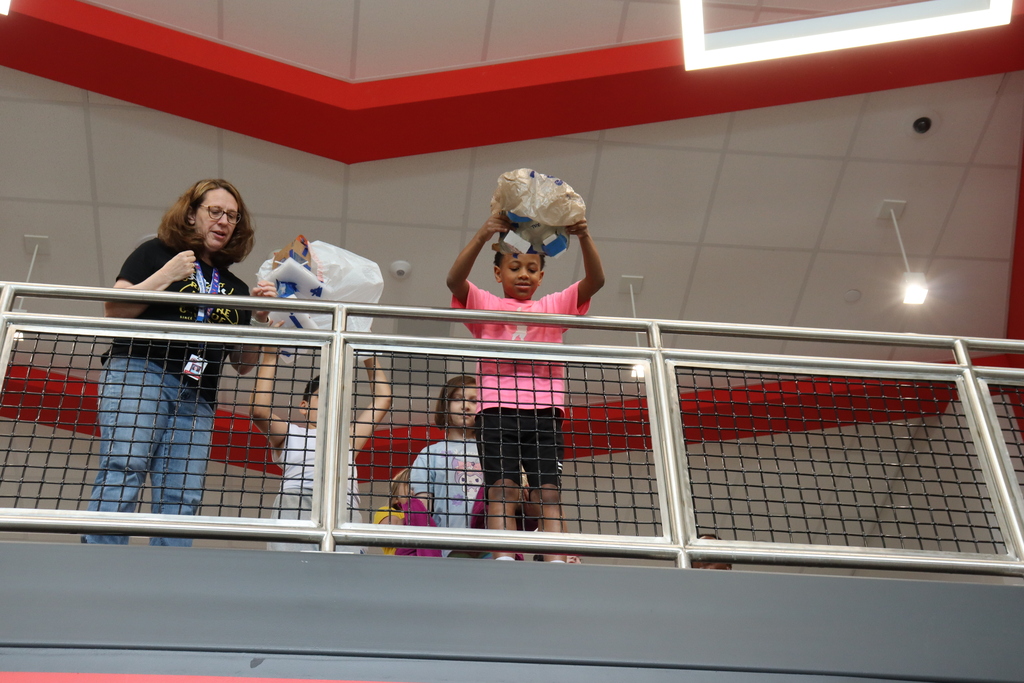 Adults and children stand on a balcony with railings. Some hold bags. The ceiling has a red line.