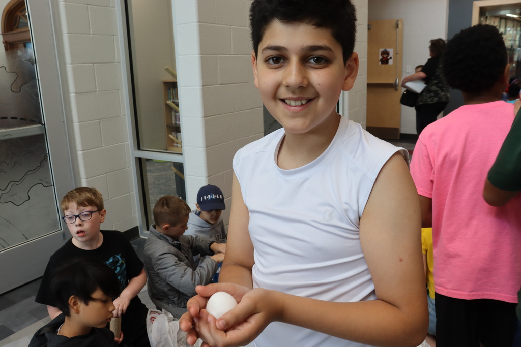 A smiling boy holds an egg and poses indoors. Other children sit in the background.