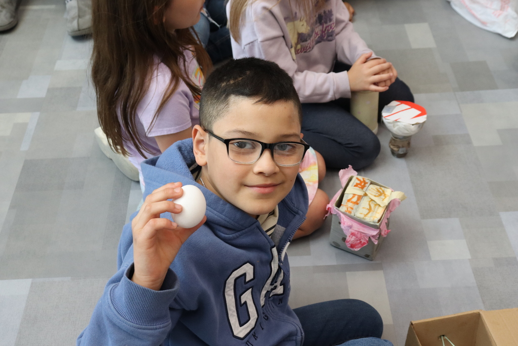 A young boy in a blue hoodie holds a white egg while sitting on a patterned floor, surrounded by other children.