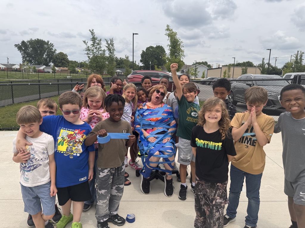 A group of children posing for a photo on a concrete sidewalk, with a fence, trees, and parked cars in the background and their teacher taped to a chair