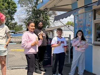 Five people stand in front of an ice cream stand, each holding a cone. Trees and a fence are in the background.