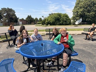 A group of individuals are seated around a blue table in an outdoor area with benches, trees, and a road in the background.