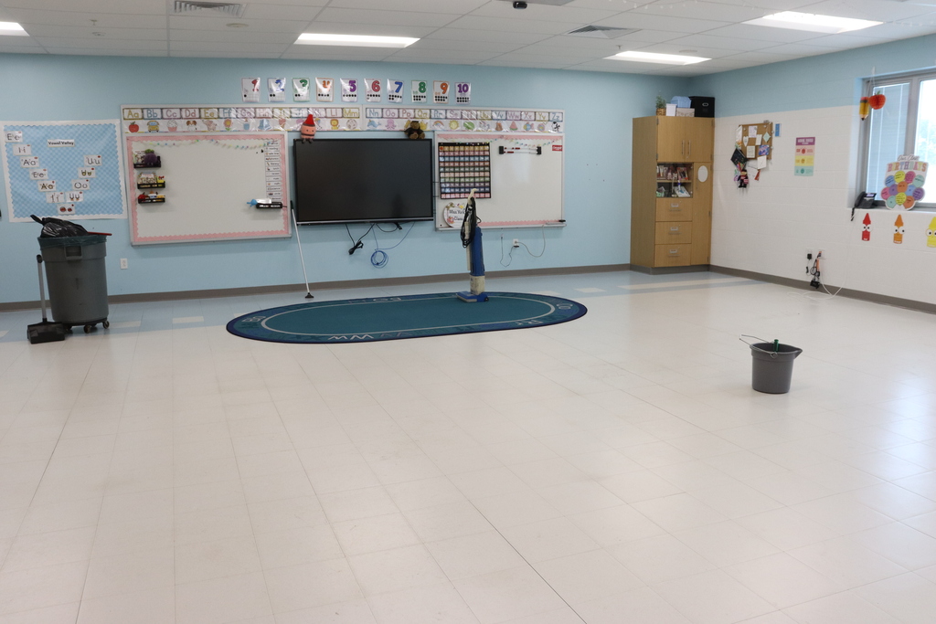 Classroom with white tile floor, blue walls, trash bin, and rug. A television and whiteboard are on the wall.