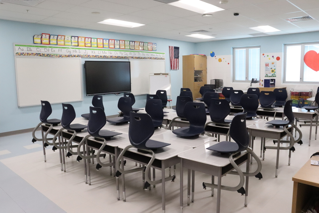 An empty classroom with desks, chairs, whiteboards, a TV, and a US flag on the wall.