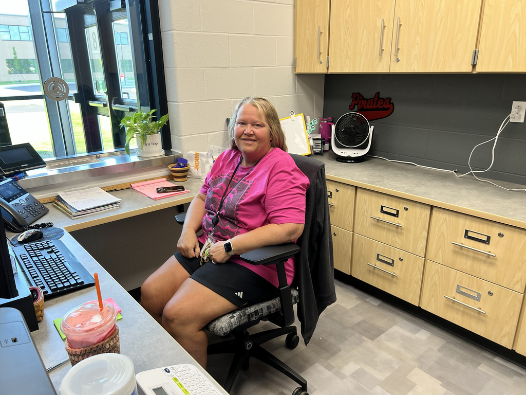 A woman sits at a desk in an office, wearing a pink shirt and black shorts. A cup sits on the desk.