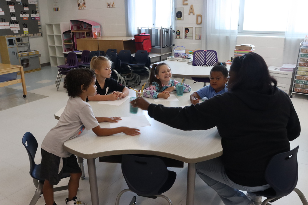 A classroom with white walls. A woman sits at a table with four children. One child sits at the edge of the table.