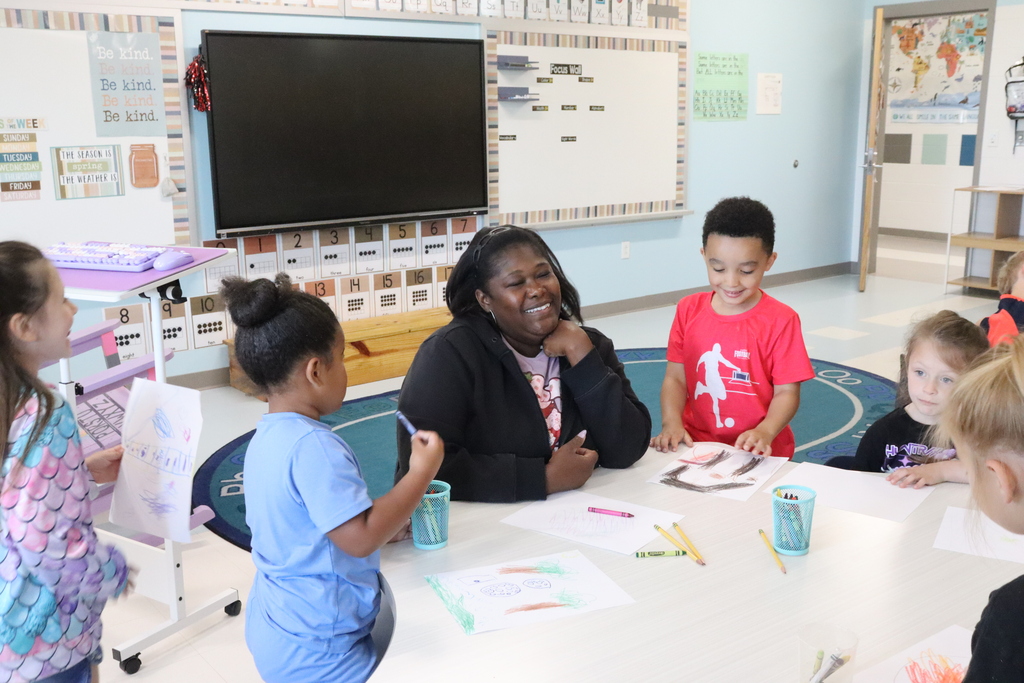 Classroom with children and an adult. They sit around a table with art supplies. A large screen is behind them.