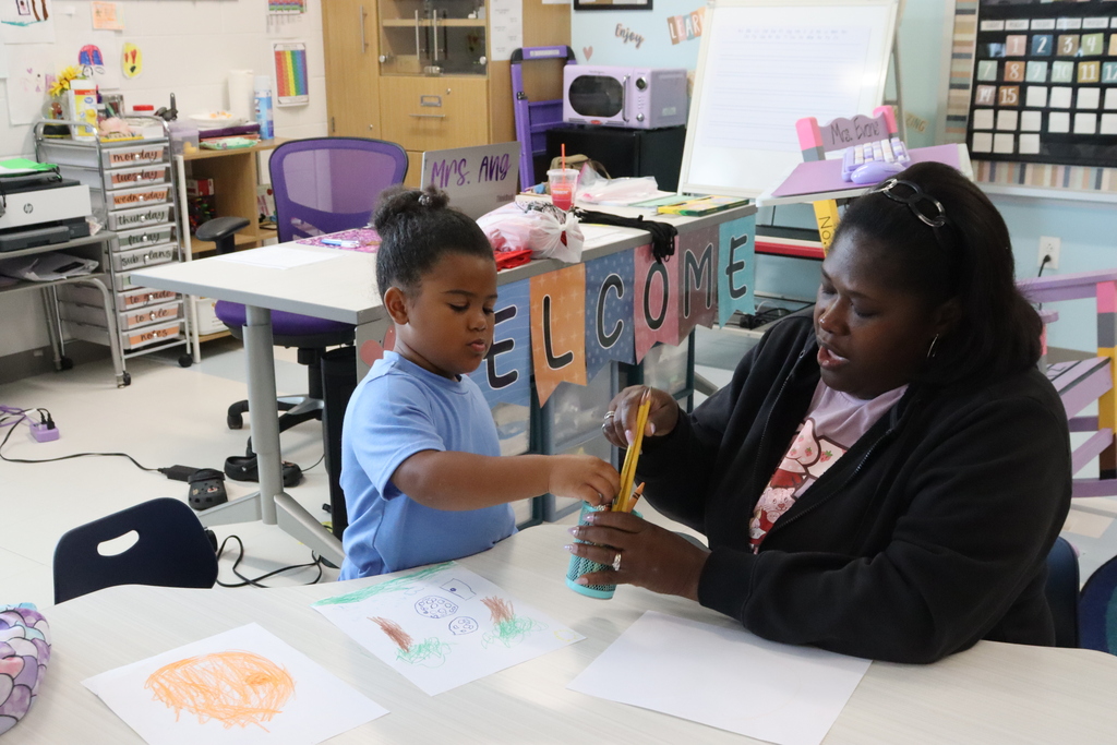 A teacher and child are seated at a table. They are both using art supplies.
