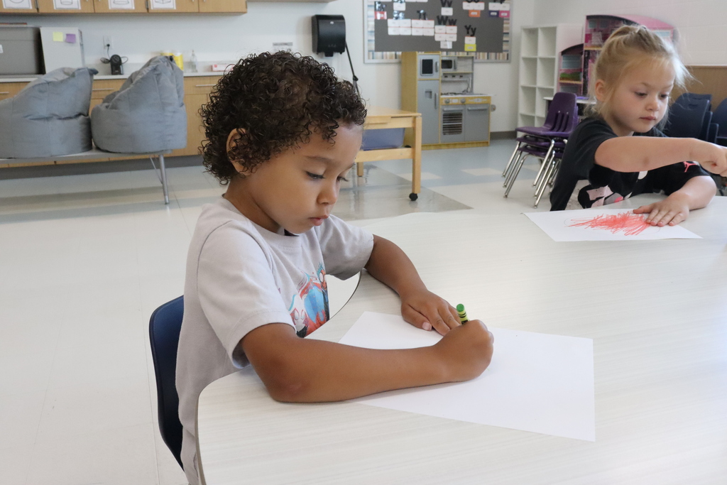 Two young children are seated at tables in a classroom. One child has curly hair and is coloring with a crayon.