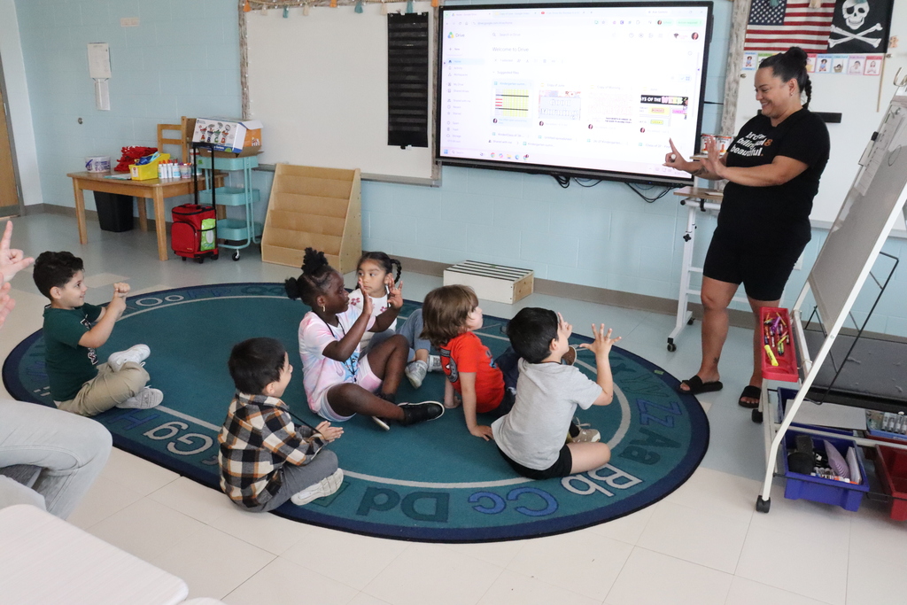 A classroom with a teacher presenting on a screen while children sit in a circle on the rug.