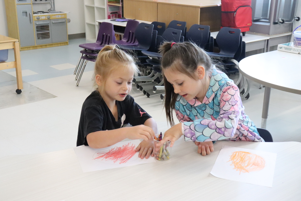 Two girls seated at a table in a classroom, each working on an art project, surrounded by chairs.
