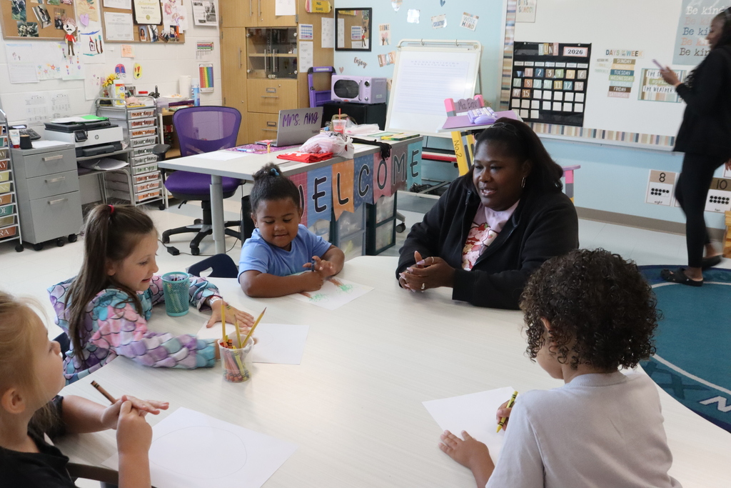 A woman talks with four children seated around a table in a classroom. Each child has a paper and pencil.