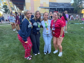 Four women in varied outfits, standing close together on a grassy area. A building with a multicolored facade in the background.