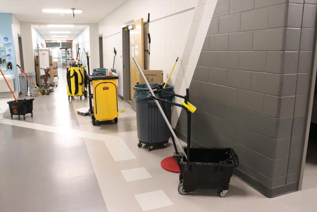 An empty hallway with cleaning equipment, including a yellow bucket and black and grey push brooms, lined up against a brick wall.