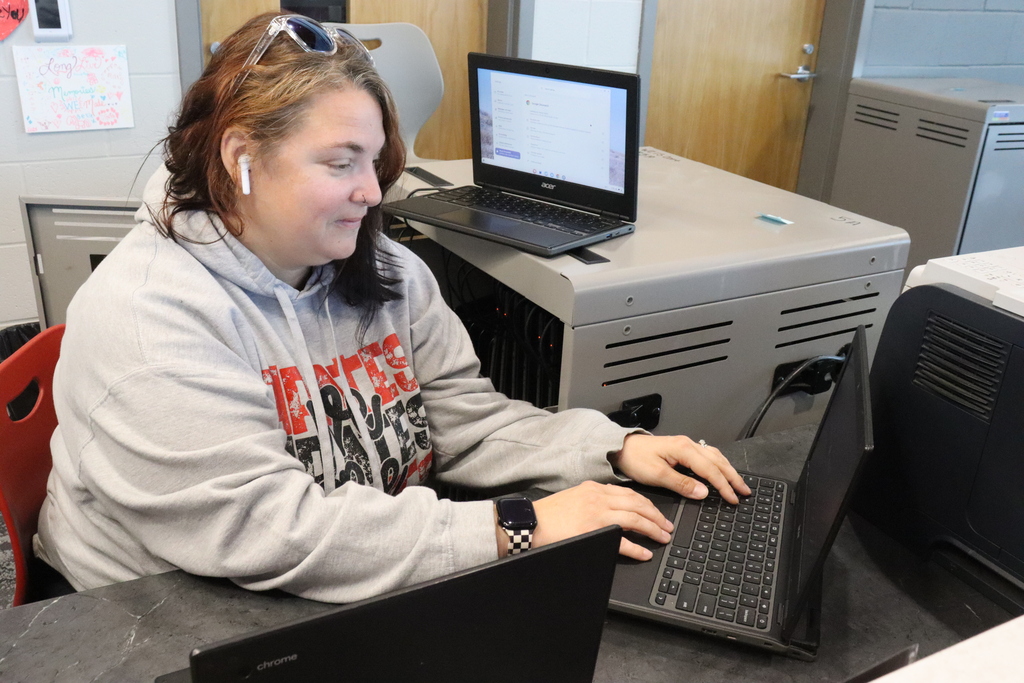 A woman in a gray hoodie sits at a desk with two laptops, one open, and earphones in her ears.