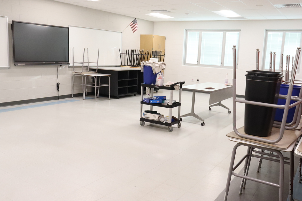 Empty classroom with white walls, windows, a large screen, chairs, and desks. Trash bins and a trolley near the center.