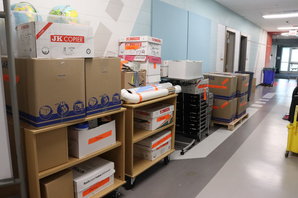 Boxes and storage containers stacked on shelves and trolleys in a hallway with white walls.