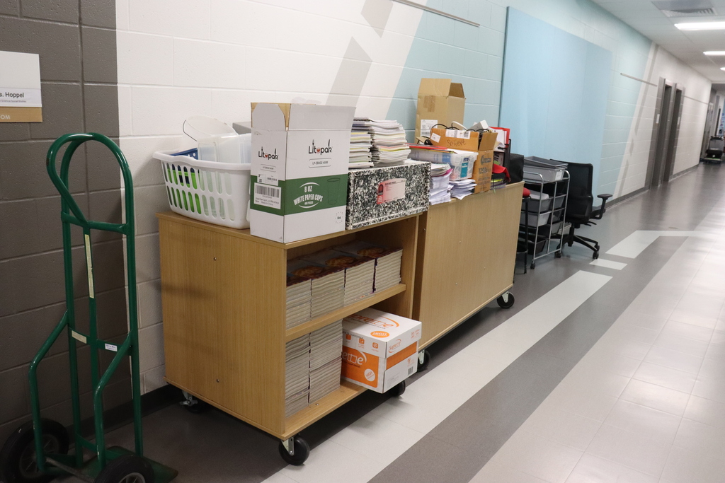 A hallway with shelves carrying boxes and containers. A dolly stands to the left. The floor has stripes.