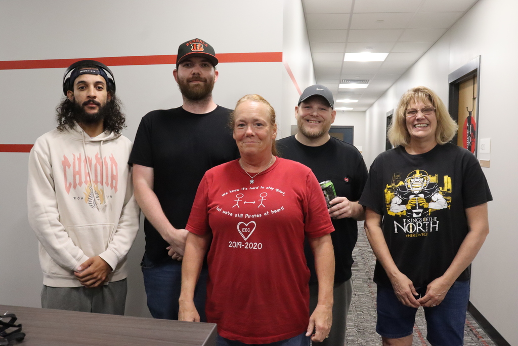 Five people stand together in a hallway; one wears a red shirt, one a white hoodie, one a black shirt, and two wear black tees.
