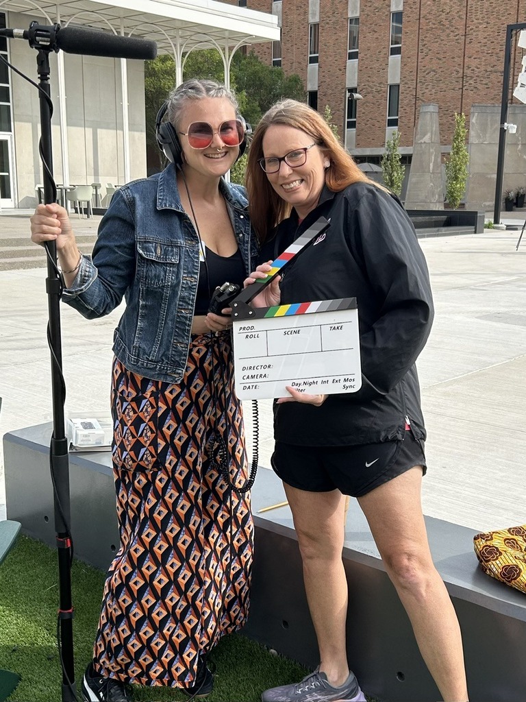 Two women stand on a grassy area holding a clapperboard. One wears a denim jacket, the other a black jacket. Both wear glasses. A building and greenery are in the background.