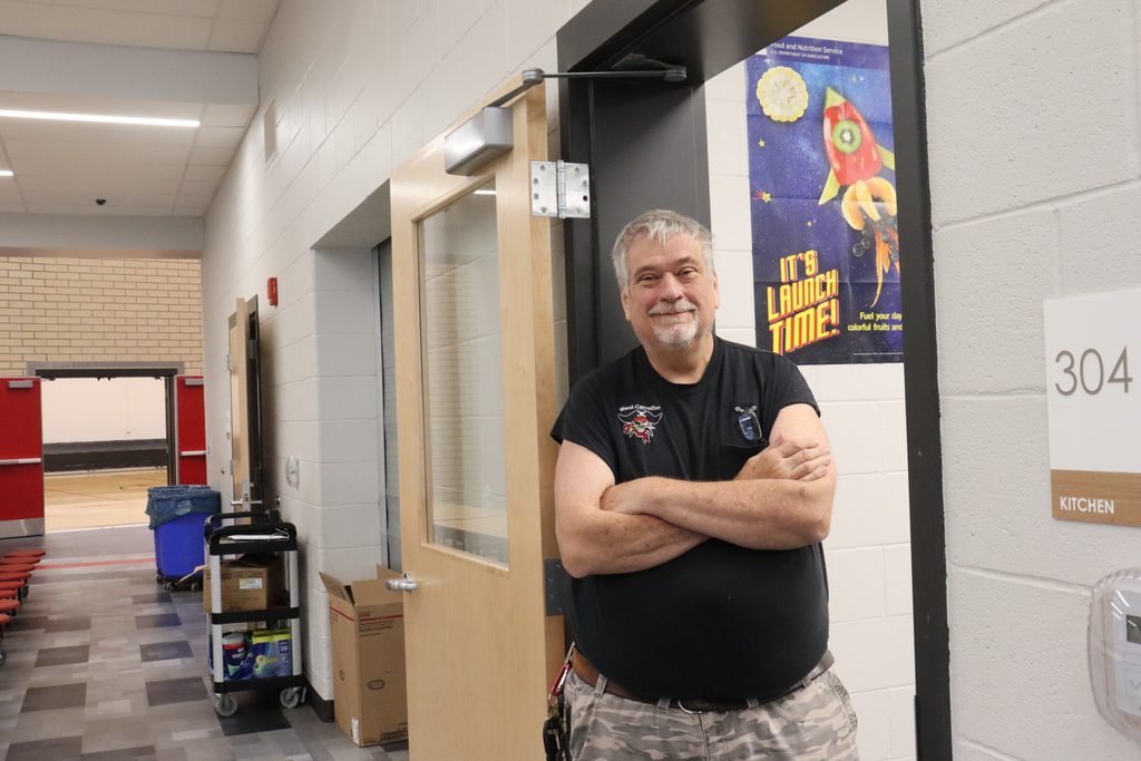 Man in a black t-shirt and pants stands with arms crossed in a hallway. Behind him, a door and a poster are visible.