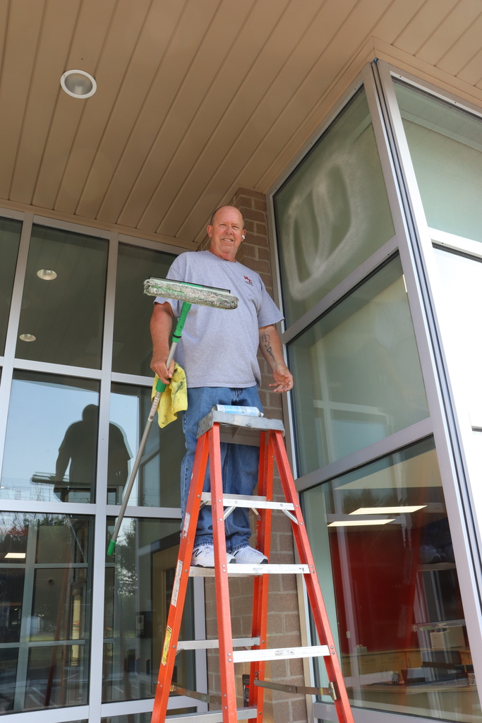 A man is standing on a ladder cleaning a glass door. He is wearing a blue shirt and blue jeans.