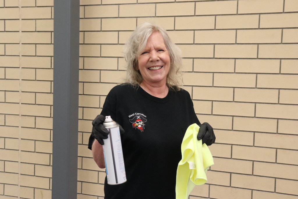 A woman in a black T-shirt smiles and holds a white spray can and yellow cloth.