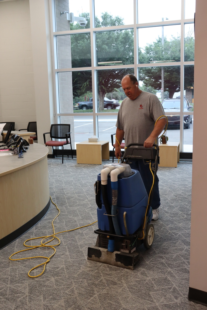 A man wearing a gray shirt is using a carpet cleaner in a room with a large window.