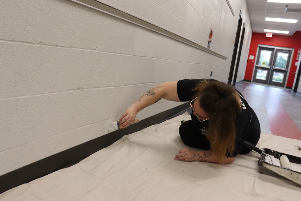 A person in black clothing kneels on a white floor, applying paint to a wall with a trim brush.