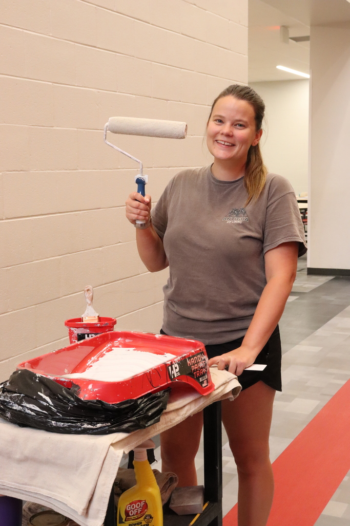A woman holds a paint roller, standing behind a table with a red tray and paint supplies.