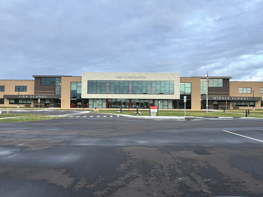 A school building with large windows and a sign reading "West Carrollton". An empty parking lot in front.