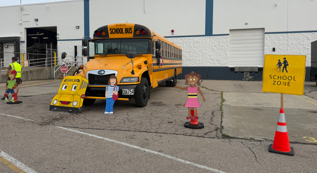 School bus in front of building. Two figures stand next to it. A yellow sign reads "SCHOOL ZONE" next to a traffic cone.